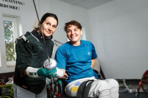 Two women fencers smile during a wheelchair fencing session indoors, showcasing adaptive sports.