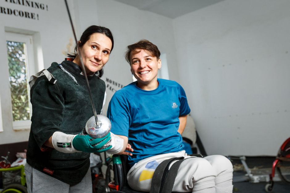 Two women fencers smile during a wheelchair fencing session indoors, showcasing adaptive sports.