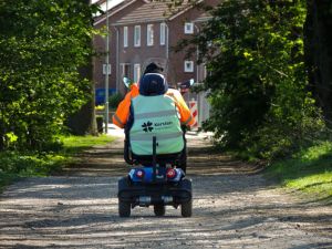 An adult riding a mobility scooter along a tree-lined path near residential buildings.