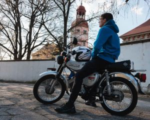A man in a blue jacket sits on a Simson motorcycle by a white wall and historic clock tower.