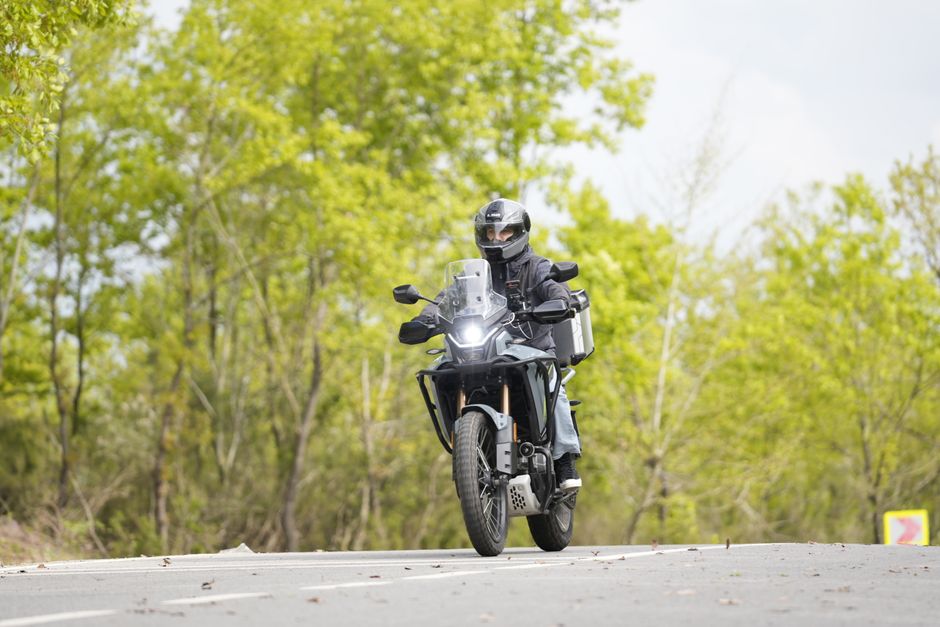 A motorcyclist in safety gear rides through a scenic forest road during daytime.