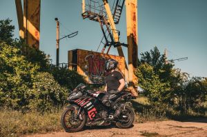 A biker poses with a motorcycle in an outdoor industrial area under bright sunlight.